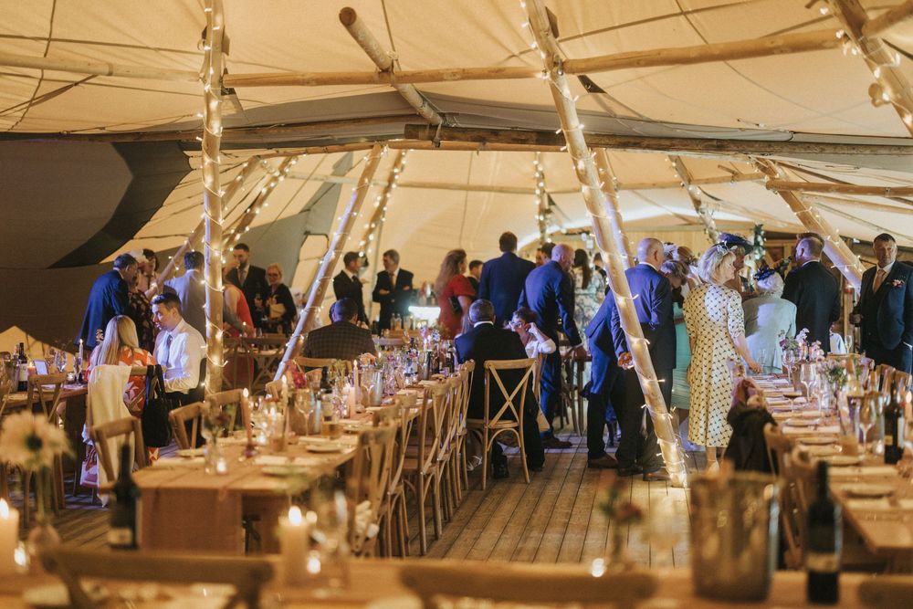 Wedding reception guests dine under a warm-lit tent with rustic wooden chairs and elegant table settings at Scrivelsby Walled Garden.