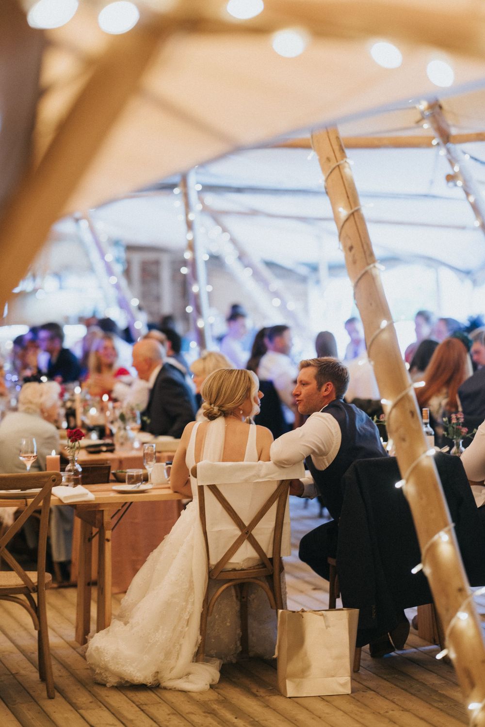 Newlyweds seated at rustic wooden table under string lights in reception tent during wedding celebration at Scrivelsby Walled Garden.