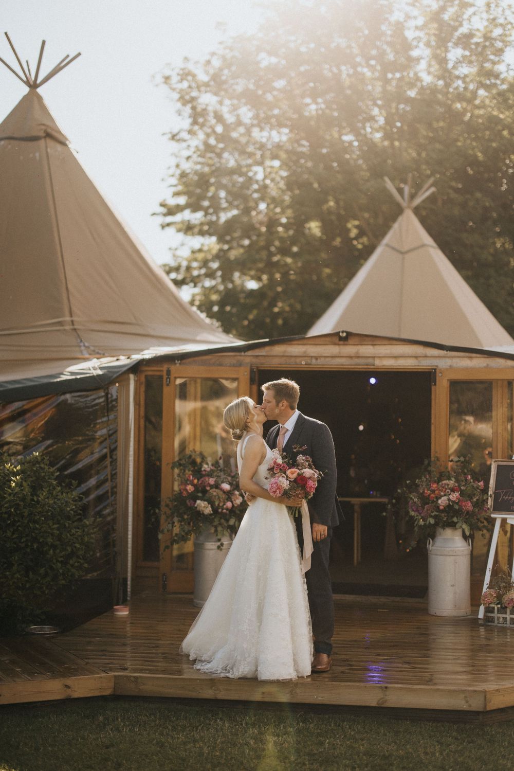 Newlyweds share a romantic moment on a wooden deck in front of two rustic tipi tents at sunset at Scrivelsby Walled Garden.