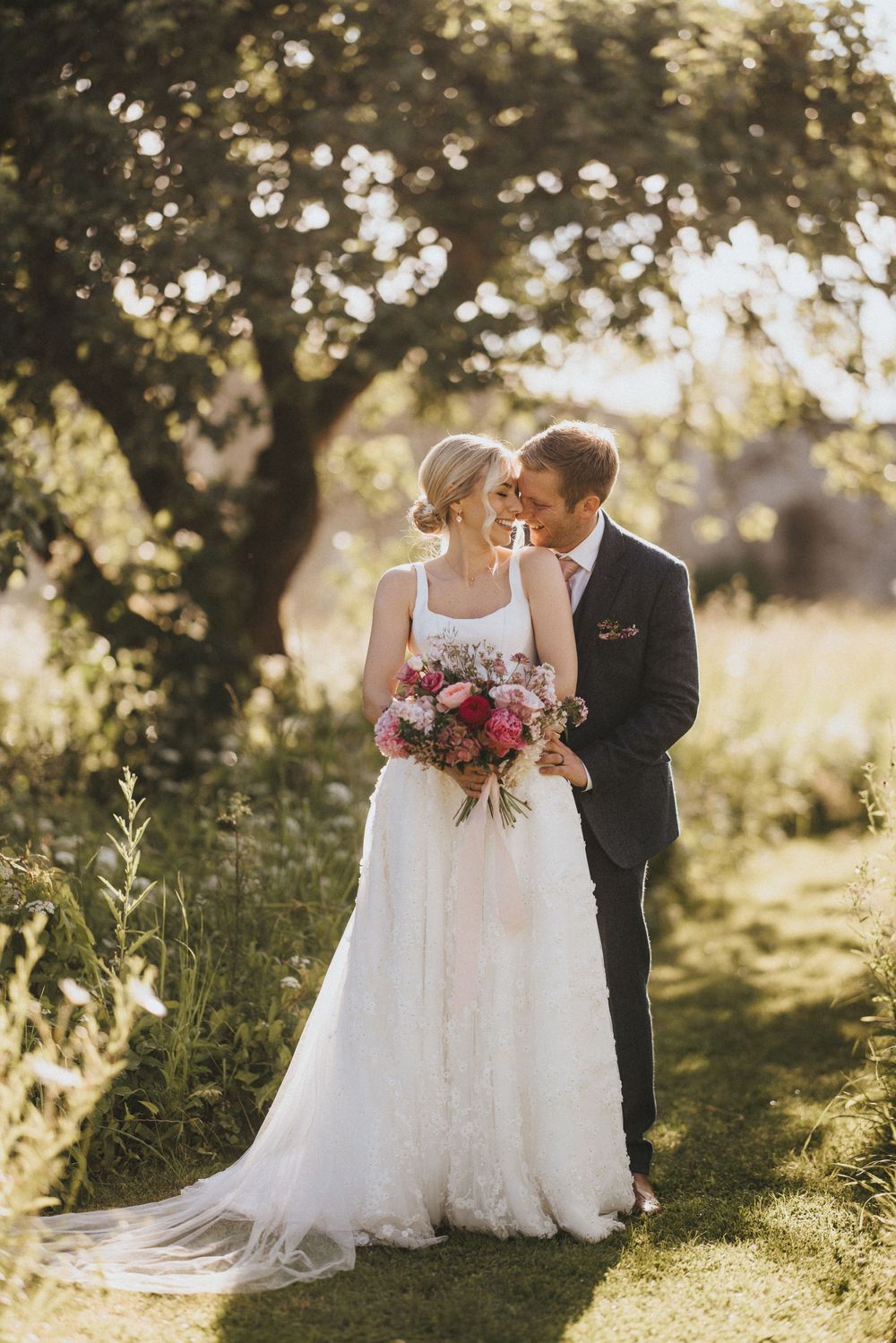 A romantic wedding couple embraces in a sun-dappled garden setting with a beautiful bouquet of pink flowers at Scrivelsby Walled Garden.