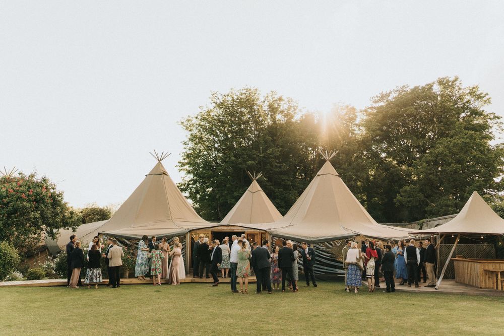 Several tipi-style event tents stand in a row on a grassy field during golden hour as guests gather outside at Scrivelsby Walled Garden.