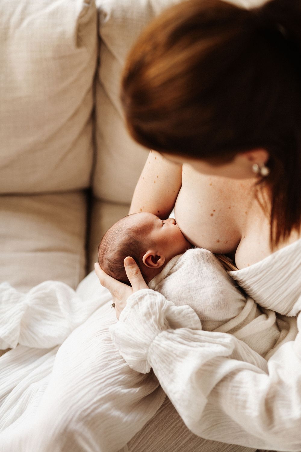 Intimate moment of mother nursing newborn baby wrapped in soft white fabric on couch.