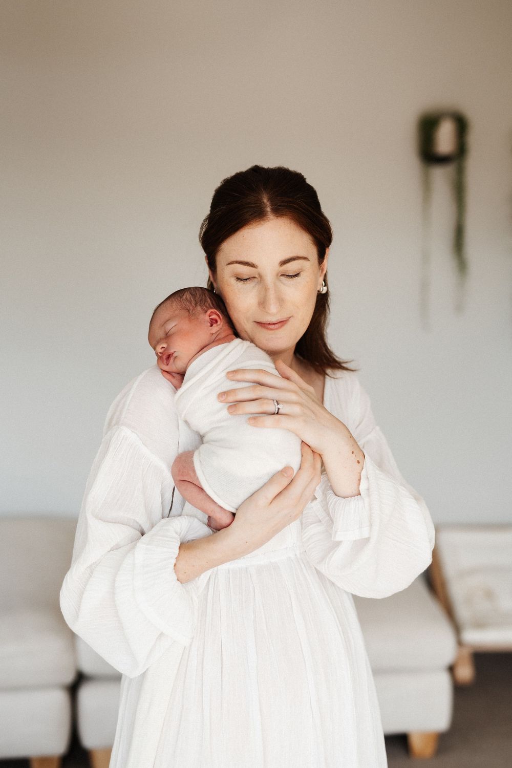 A tender moment captured as a woman in a flowing white dress holds a sleeping newborn against her chest in a minimalist room.