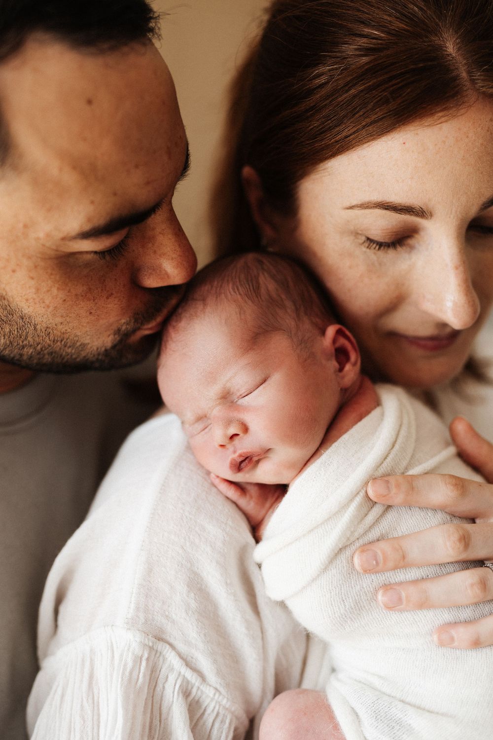 Parents snuggling close to their sleeping newborn baby swaddled in white.