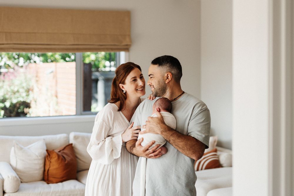 Happy parents in white clothing holding their newborn baby in bright living room.