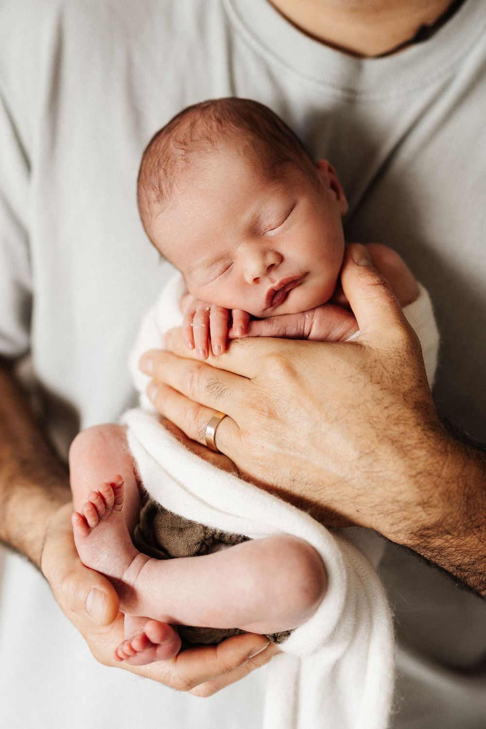 Newborn baby sleeping peacefully cradled in an adult's hands against white blanket background.