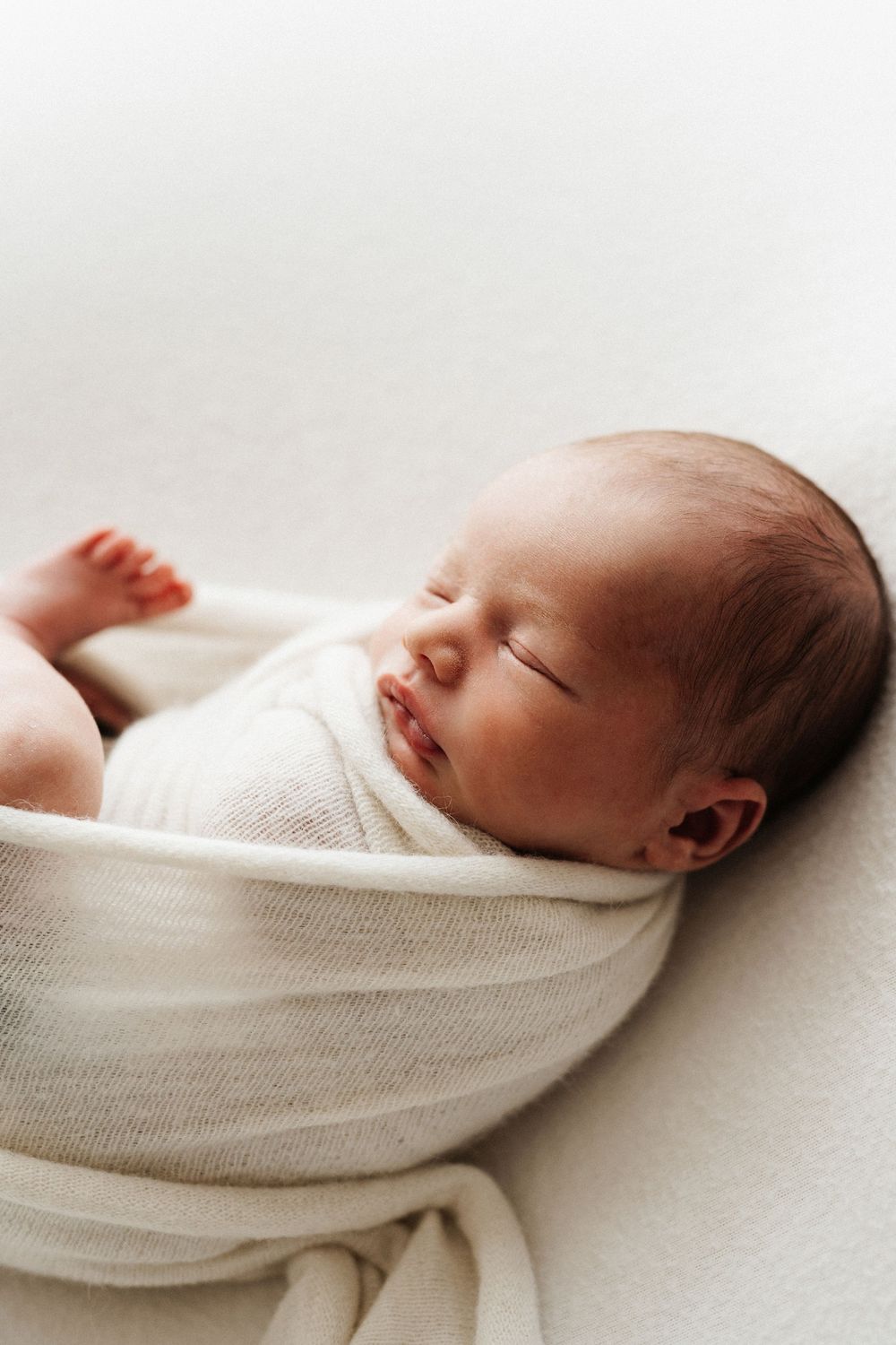 Peaceful sleeping newborn baby swaddled in soft white fabric against light background.