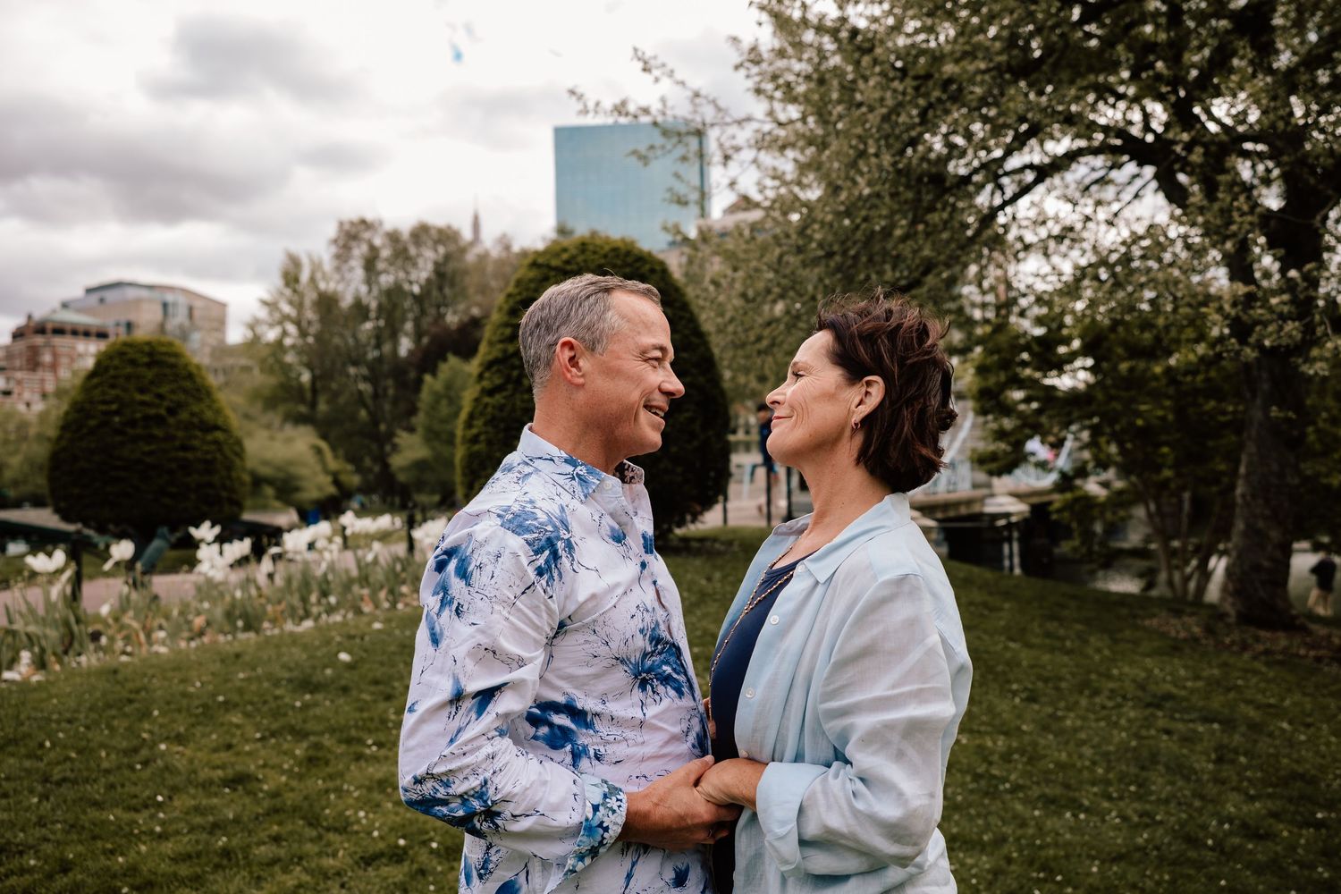 A joyful couple wearing tie-dye and light blue sweaters share a laugh together in a park with city buildings in background.