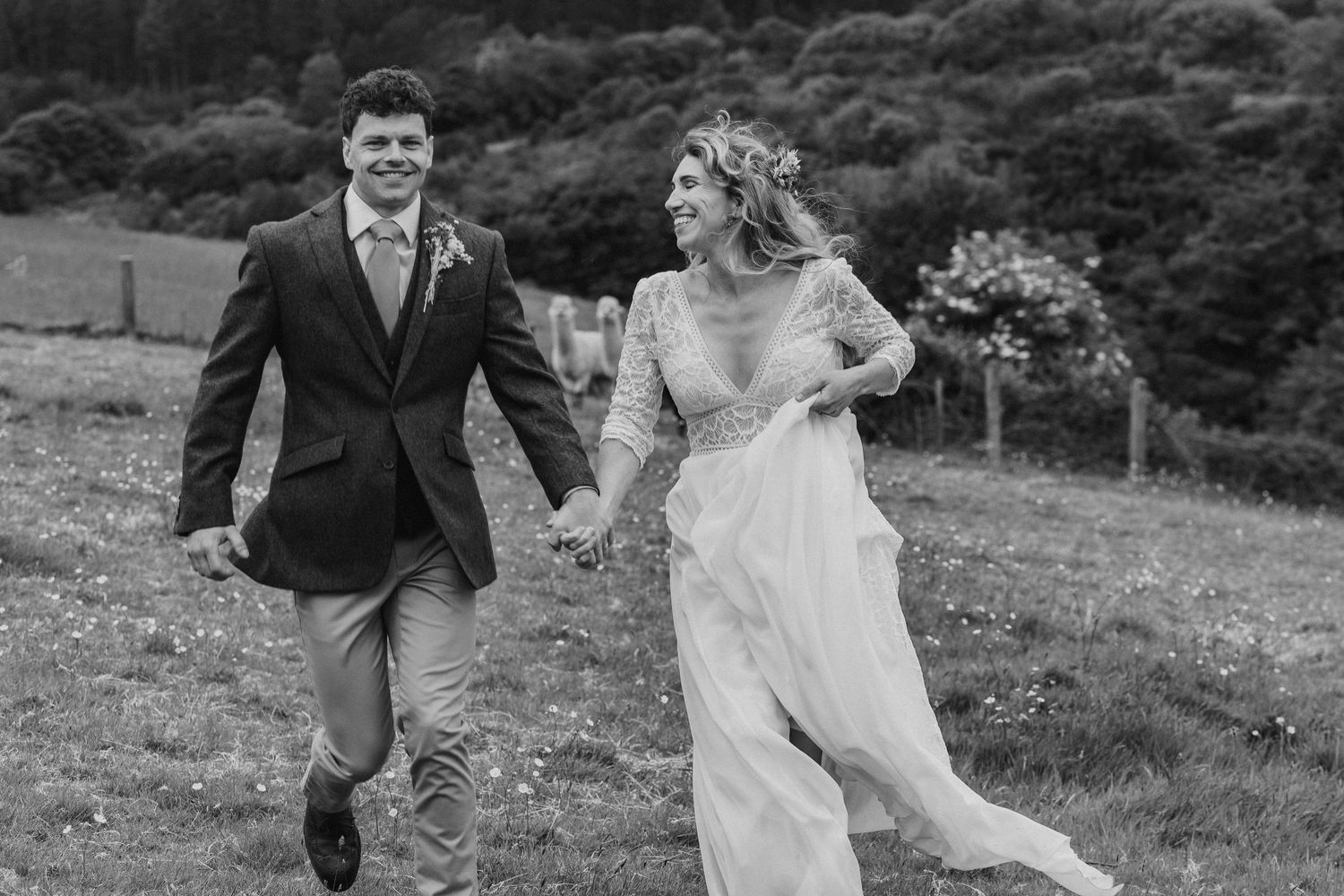 Black and white photo of newlyweds joyfully running through a hillside meadow holding hands.