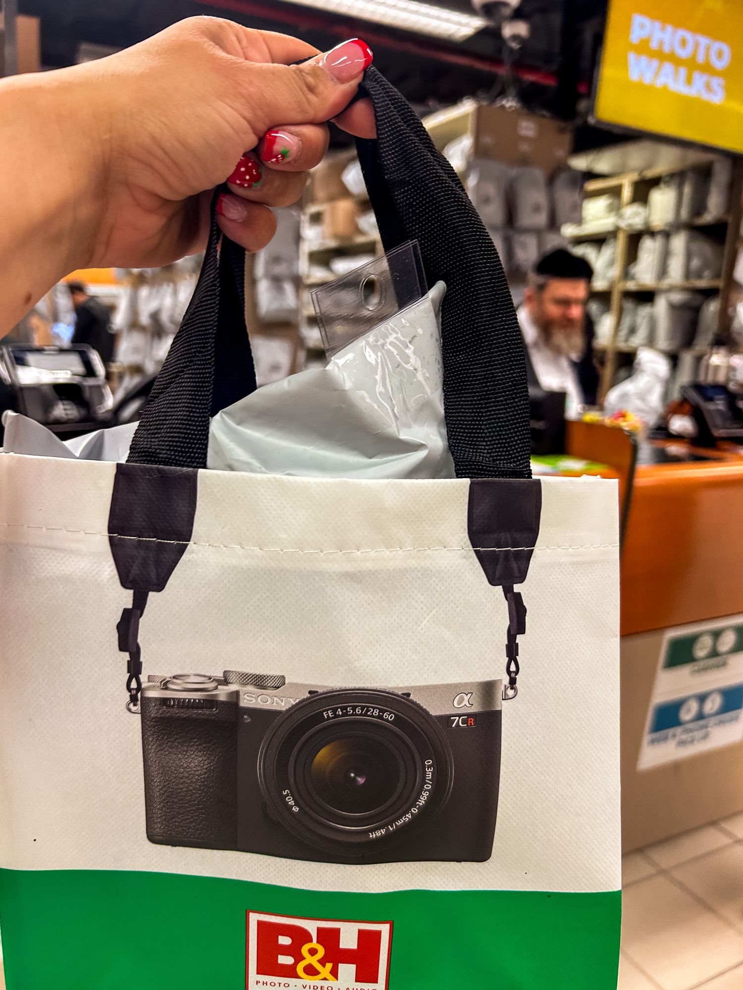 A white shopping bag from B&H Photo featuring a printed camera design with black handles being held in a retail store.