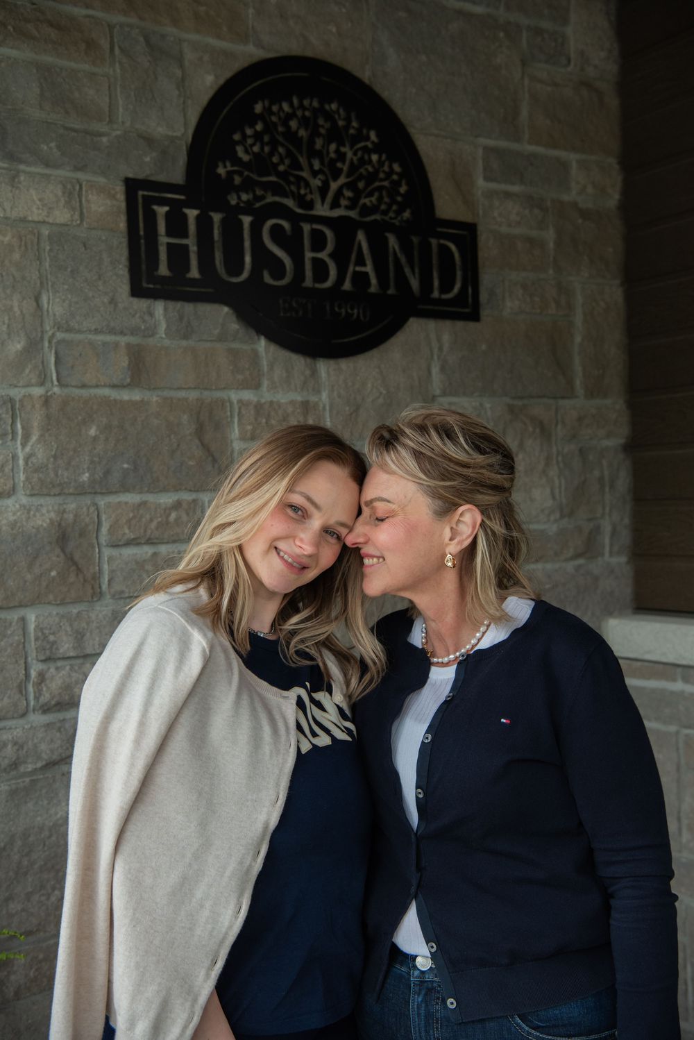 Two people share intimate moments while leaning against stone wall with 'Husband' sign visible.