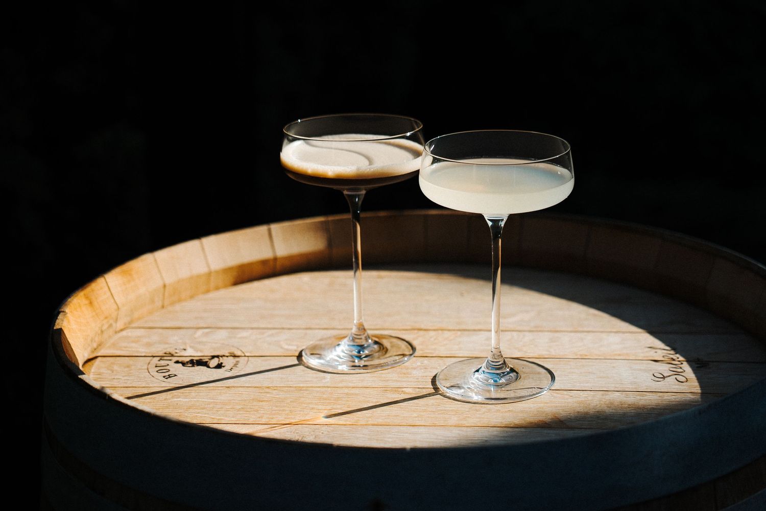 Two elegant cocktail glasses sit atop a curved wooden surface against a dark background.
