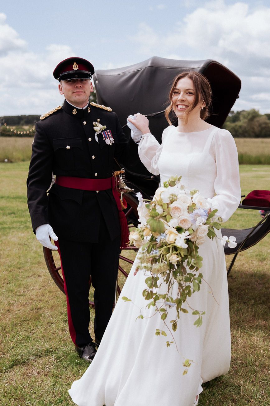 Bride in elegant long-sleeved white gown holds cascading floral bouquet next to military officer in formal uniform outdoors.