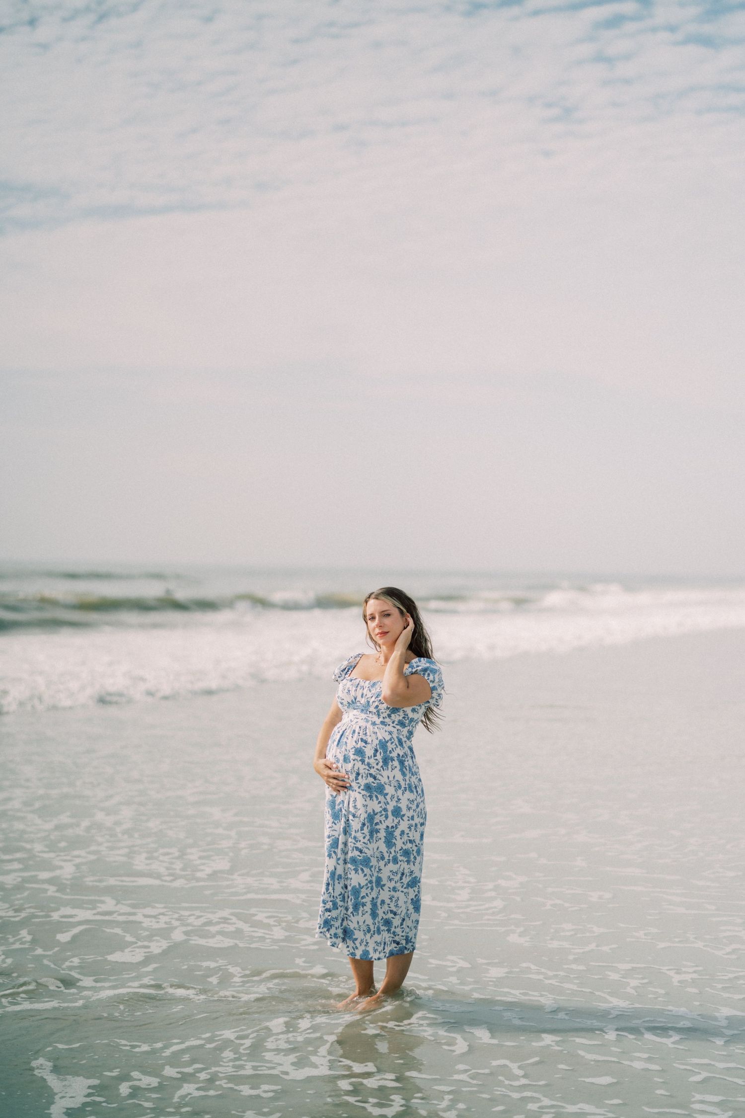 Person in a blue floral dress stands in shallow ocean waves on a cloudy day at the beach.