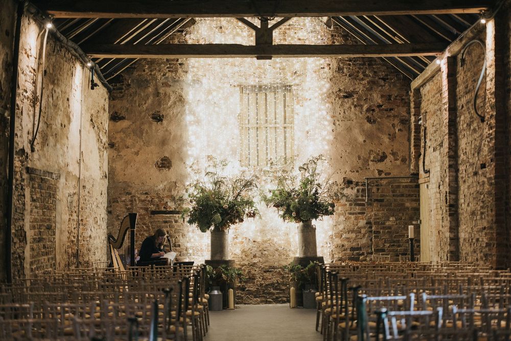 Interior of rustic stone chapel with wooden chairs and two floral arrangements in front of weathered stone walls.