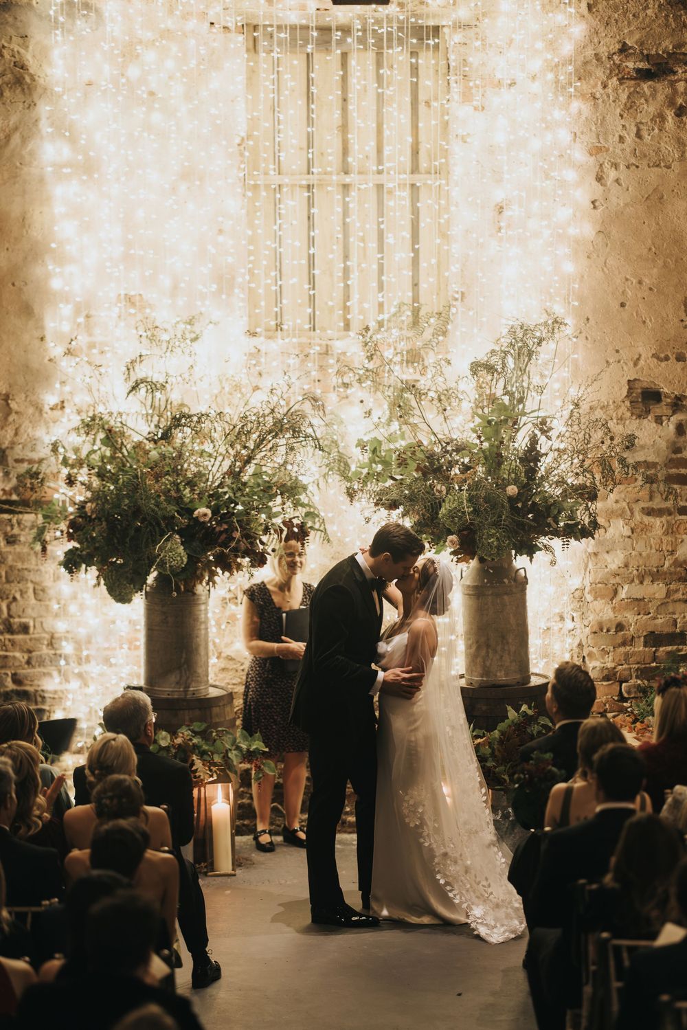 A romantic wedding ceremony takes place in a rustic brick venue decorated with greenery and soft lighting.