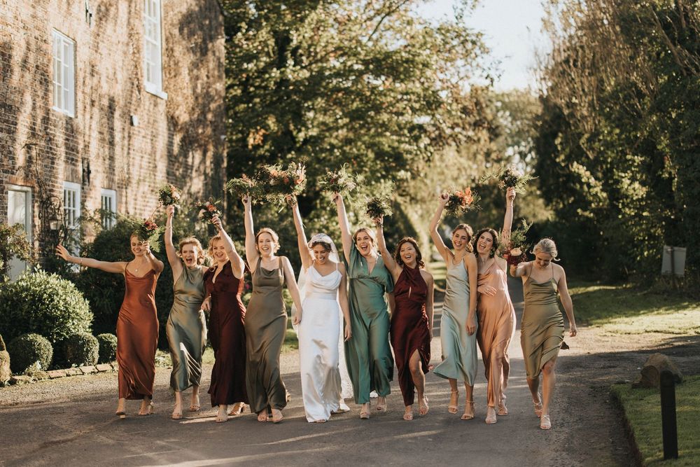 Bridesmaids in mix-matched earth tone dresses walk arm in arm down a sunlit garden path lined with trees.