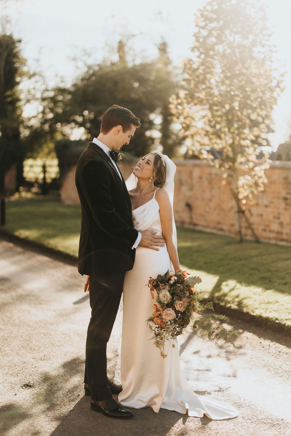 Romantic couple in wedding attire embrace at golden hour holding autumn bouquet along garden path.