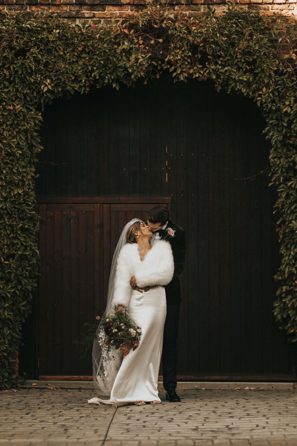 A couple in wedding attire embrace in front of a rustic wooden door framed by elegant green foliage.