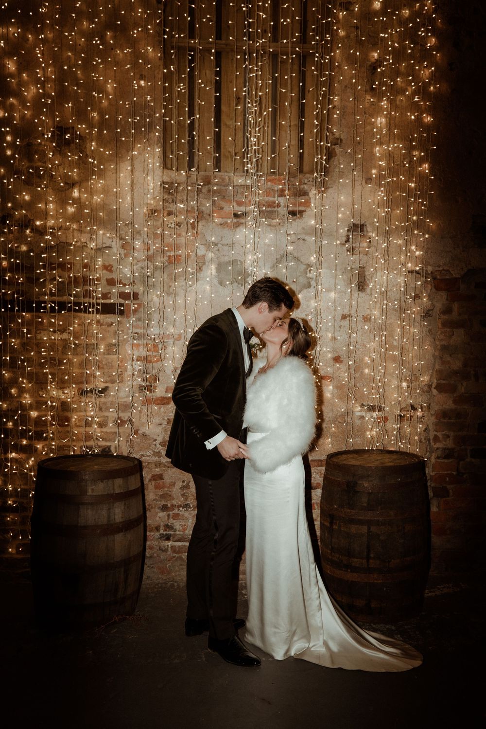 A romantic couple embraces between rustic wine barrels under a magical backdrop of twinkling string lights.
