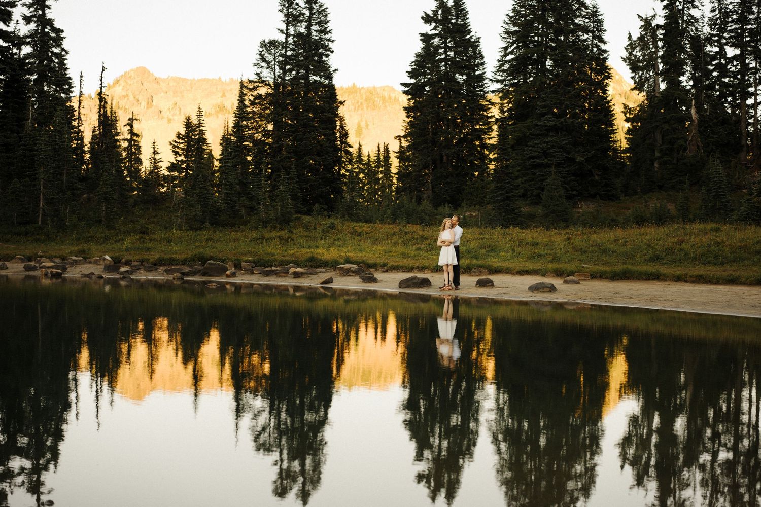 A silhouetted figure stands by a tranquil mountain lake at sunset with evergreen trees reflecting in the still water.