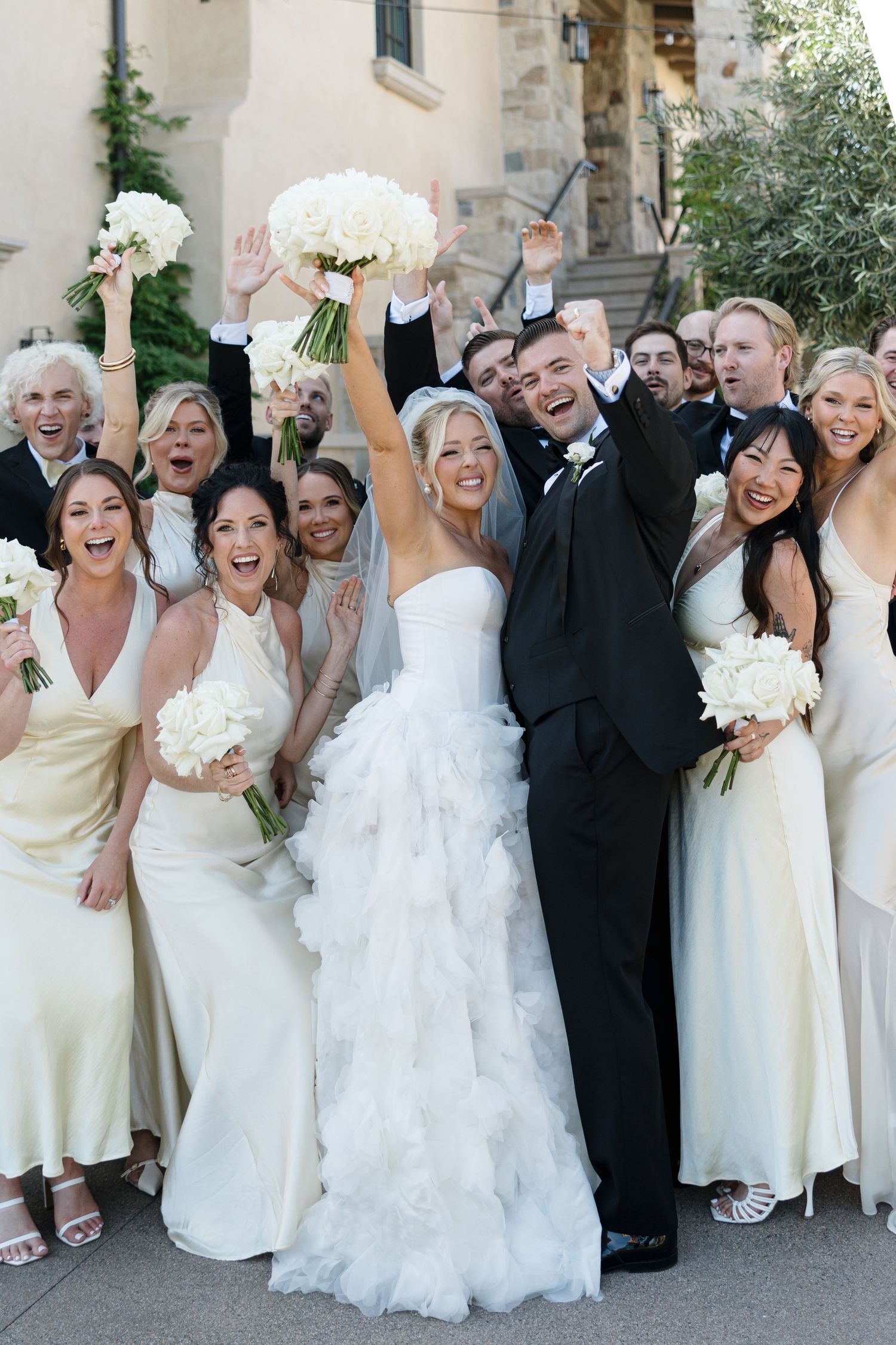 A wedding party in formal attire poses together while celebrating and cheering on outdoor stone steps.
