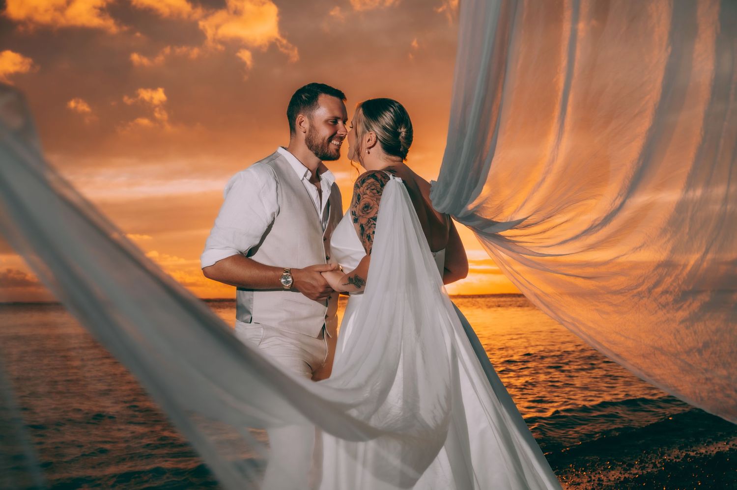 Happy newlyweds walking on tropical beach with palm trees as bride holds white bouquet on sunny day.
