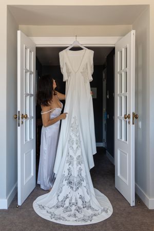 An elegant white wedding dress with intricate lace detailing hangs in a hallway against white double doors.