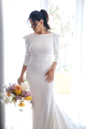 Simple white wedding dress shown with colorful floral bouquet near bright window.
