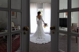 Elegant wedding dress silhouette viewed through French doors with flowing train against bright window.
