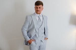 A groom in a light gray suit stands against a white wall.