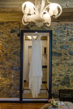 White wedding dress displayed on hangers against rustic stone wall with elegant chandelier lighting.