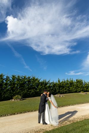 A wedding couple stands on a pathway beneath dramatic wispy clouds and blue skies.