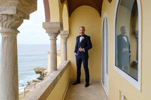 Elegant suited figure standing on Mediterranean balcony overlooking ocean view.