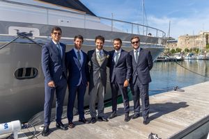 A group of men in formal suits posing together on a dock with a luxury yacht in the background.