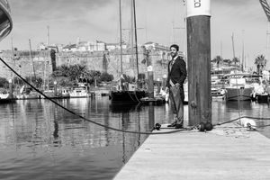 Black and white photo of a person standing on a wooden dock overlooking a marina with boats.