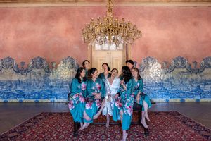 Wedding party sits in a row wearing coordinated teal robes in an elegant room with chandeliers.
