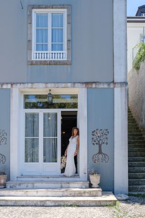 Bride in white dress photographed through doorway of light blue Mediterranean building.