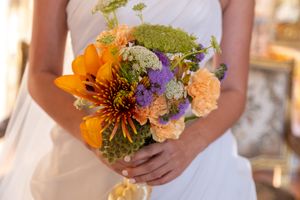 Wedding bouquet featuring bright orange lilies and purple flowers held against white dress.
