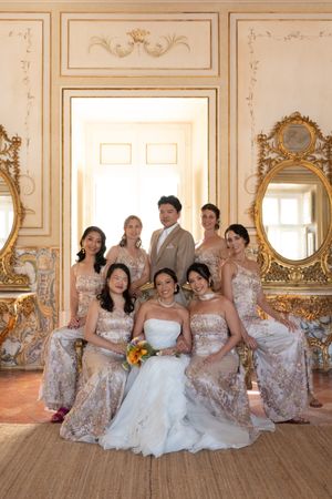 Wedding party in sparkly dresses poses together in an ornate room with gilded mirrors.