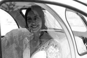 Bride sitting in classic car wearing white wedding dress and smiling in black and white photograph.