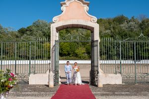 Couple stands under decorative pink archway entrance with red carpet and iron gates.