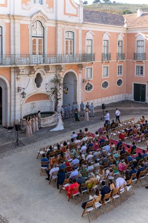 Aerial view of outdoor wedding ceremony with guests seated in rows at pink historic building.
