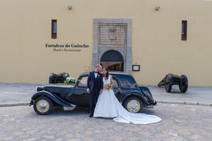 Wedding photos with vintage black car parked outside a yellow stone building.