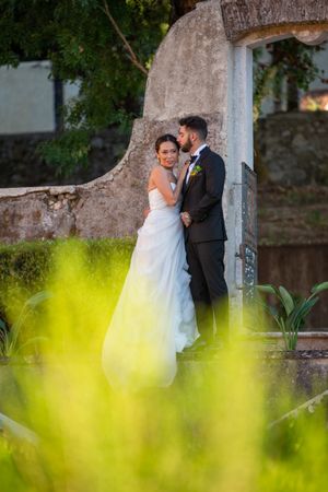 Romantic wedding photo through yellow flowers showing couple against stone wall.