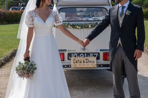 A wedding couple holds hands in front of a classic car on a sunny day.