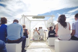 Guests watch a beach wedding ceremony under a white canopy against blue skies.