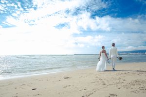 A couple in wedding attire walking hand in hand on a pristine beach with blue ocean and sky in the background.