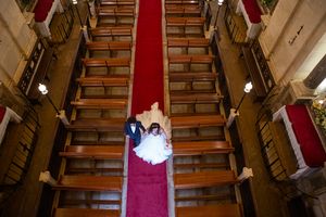 Wedding party captured from above on grand wooden staircase with red carpet.