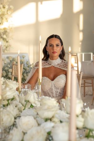 Bride seated at elegant table with white roses and tall candles in romantic lighting.